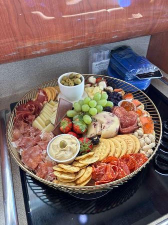 Colorful round charcuterie board on a kitchen countertop with mixed cheeses, green grapes, strawberries, olives, crackers, hummus, nuts, jam and assorted cured meats — gourmet appetizer platter.