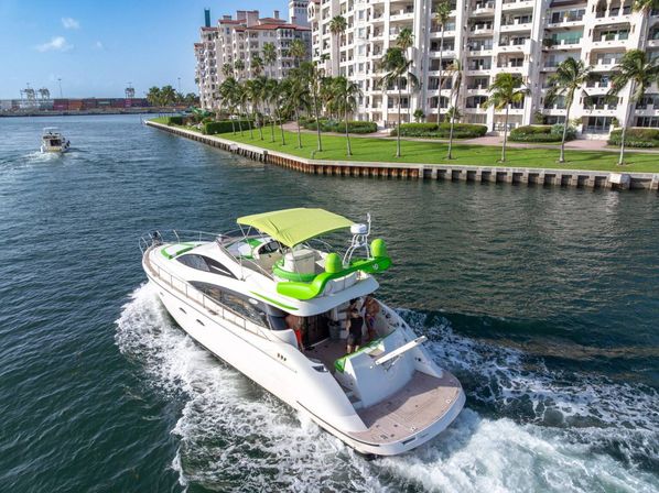 White luxury motor yacht with lime-green canopy cruising a marina channel past palm‑lined waterfront condos on a sunny coastal city waterfront, leaving a foamy wake.