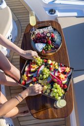 Overhead view of colorful fruit and cheese platters on a wooden yacht table — grapes, watermelon, dragon fruit, starfruit, berries, olives, sliced cheeses, crackers, two mimosa flutes, and hands reaching for fruit.