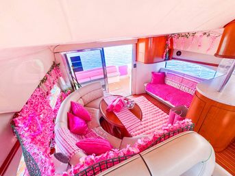 Playful pink-themed yacht interior: curved white leather lounge with bright pink pillows and throws, floral garlands and a plush rug around a wooden table, sliding glass door opening to a sunlit deck and ocean view.