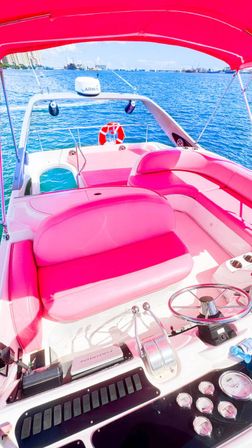 Vibrant pink speedboat interior with cushioned seating, chrome helm and steering wheel under a pink canopy, overlooking turquoise sea and distant coastal skyline on a sunny day.