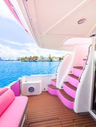 Pink-accented luxury yacht deck with teak flooring and curved stairs overlooking a turquoise bay and tropical waterfront homes under a sunny blue sky.