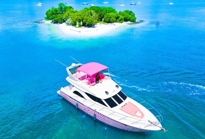 Aerial view of a pink-accented luxury yacht cruising turquoise tropical water near a small sandy island with white sand and green foliage.