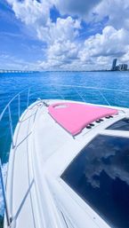 White yacht bow with pink sunpad on turquoise ocean, city skyline and distant bridge under puffy blue clouds