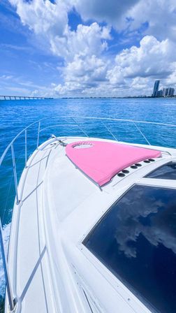 White yacht bow with pink sunpad on turquoise ocean, city skyline and distant bridge under puffy blue clouds