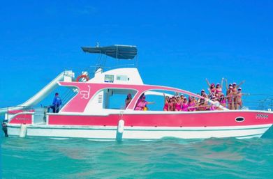 Pink-and-white party boat with waterslide cruising turquoise tropical sea, group of people in pink swimwear cheering under a clear blue sky.