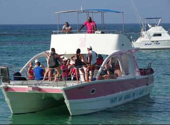 Sunlit pink-and-white catamaran party and snorkeling tour with passengers dancing and socializing on the deck above clear turquoise ocean water.