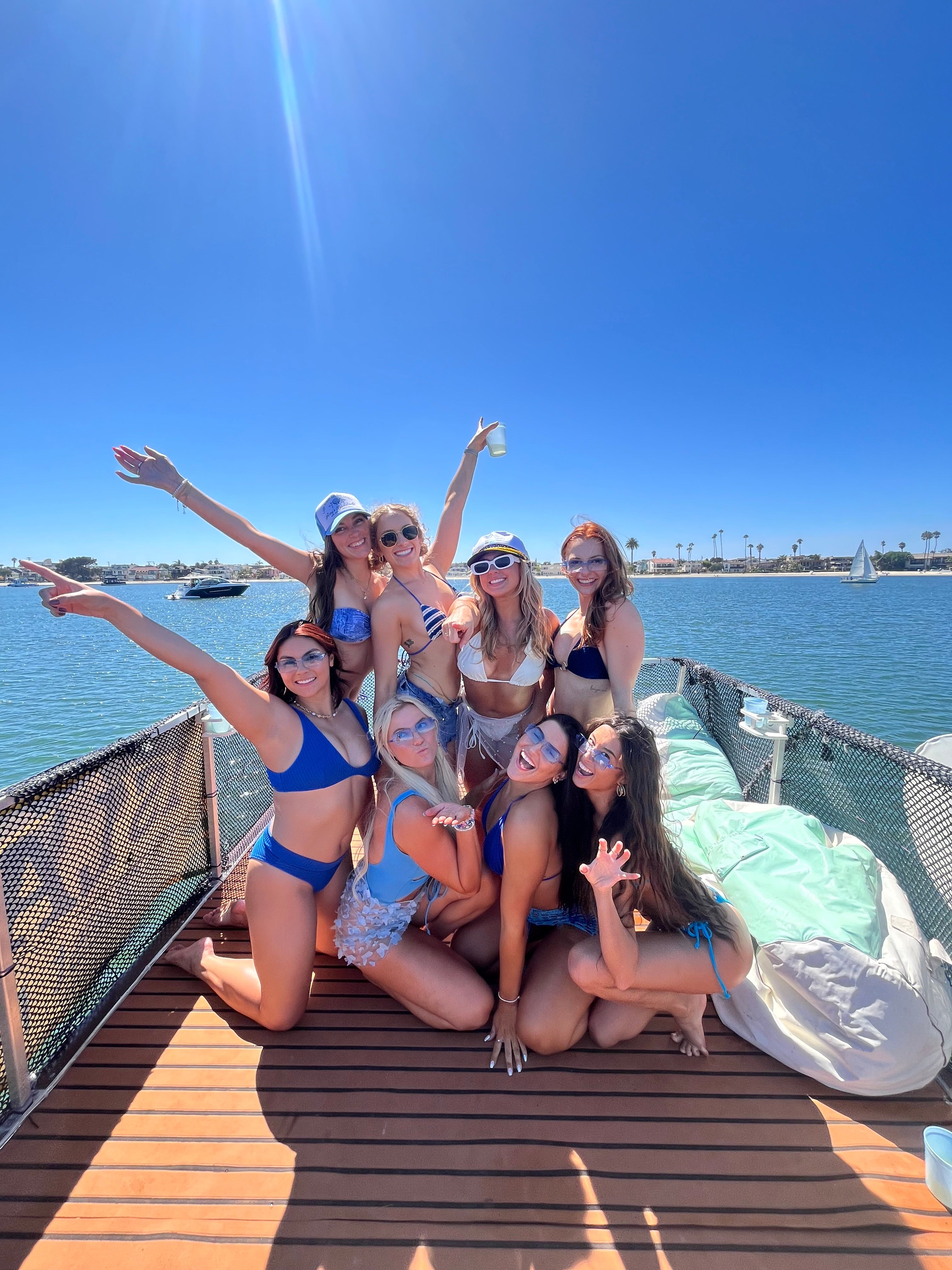Group of friends in colorful bikinis posing and laughing on a boat deck under a bright blue sky, with calm bay water, sailboats and palm trees on the distant shoreline.