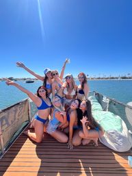 Group of friends in colorful bikinis posing and laughing on a boat deck under a bright blue sky, with calm bay water, sailboats and palm trees on the distant shoreline.