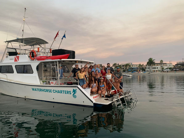 Smiling group on a charter boat at a calm marina at sunset, waterfront homes and palm trees in the background — lively coastal boat-party scene.