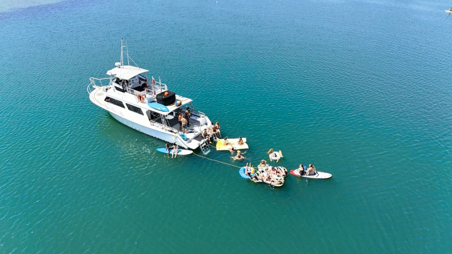 Aerial view of a white yacht anchored in clear turquoise water with people on inflatable floats, paddleboards and a swim platform enjoying a sunny day