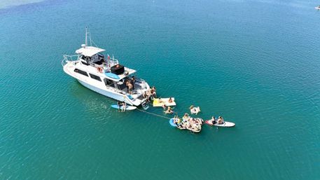 Aerial view of a white yacht anchored in clear turquoise water with people on inflatable floats, paddleboards and a swim platform enjoying a sunny day