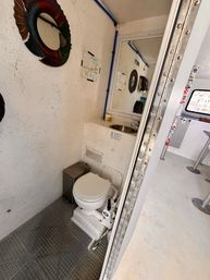 Compact onboard boat restroom (marine head) with white textured walls, manual pump toilet, stainless sink and mirror, small trash bin, decorative fish wall mirror, visible blue piping and riveted metal door frame in a vessel interior.