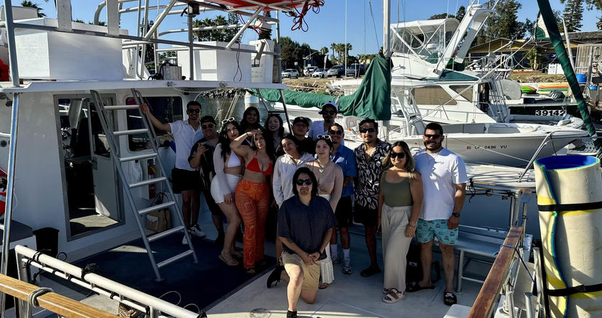 Group of friends in summer outfits posing on the deck of a sportfishing boat at a sunny coastal marina, with docked boats, palm trees and blue sky in the background.