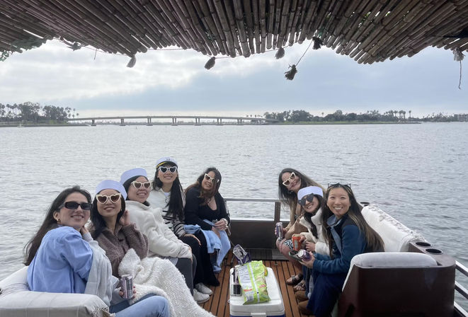 Cheerful group of friends in heart-shaped sunglasses and sailor hats lounging on a covered leisure boat, cruising a palm-lined coastal bay with a low bridge under a cloudy sky