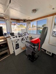 Wheelhouse of a motorboat with a gray captain's chair draped in a red life jacket, instrument-packed helm and steering wheel, and large windows overlooking a sunny marina and waterfront buildings.