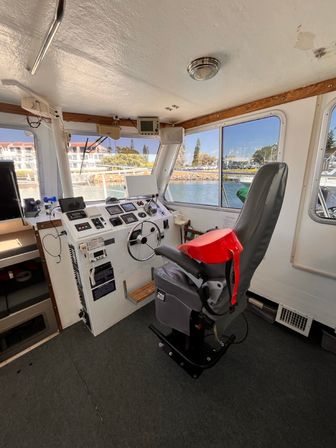 Wheelhouse of a motorboat with a gray captain's chair draped in a red life jacket, instrument-packed helm and steering wheel, and large windows overlooking a sunny marina and waterfront buildings.