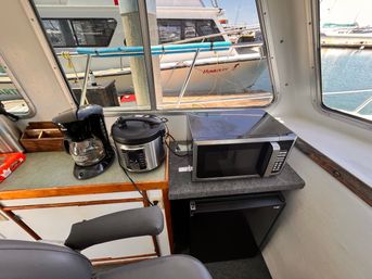 Compact boat galley with microwave, mini fridge, coffee maker and multi-cooker on a countertop, sunny marina view through windows.