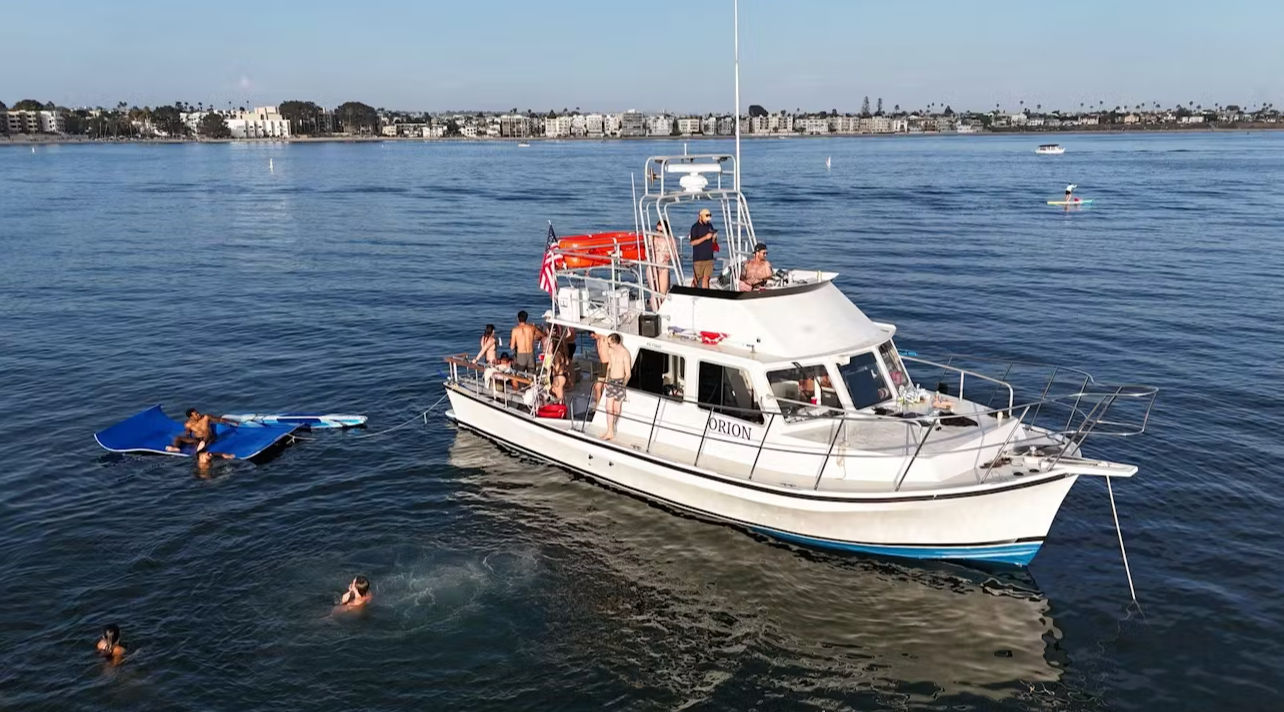 White motor yacht flying an American flag anchored in a calm coastal bay with people on deck, swimmers and a paddleboarder near a blue floating mat, with beachfront buildings on the horizon.
