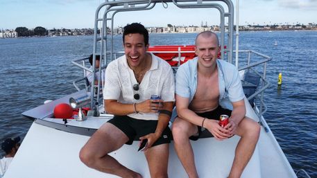 Two young men laughing on the bow of a small boat in a sunny harbor, each holding a canned drink with a coastal skyline and calm blue water behind them.