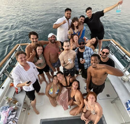 Aerial shot of a lively group of friends on a yacht party in swimsuits, smiling and holding drinks on calm open ocean water.