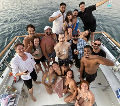 Aerial shot of a lively group of friends on a yacht party in swimsuits, smiling and holding drinks on calm open ocean water.