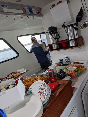 Boat party snack buffet on a small vessel: trays of chips, veggies, cupcakes and coffee urns on a counter, two people chatting by windows with a harbor/ocean view