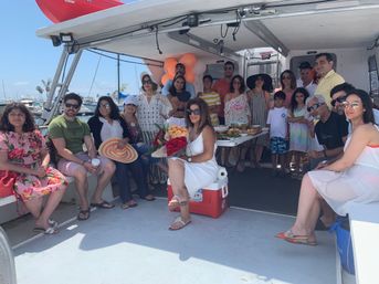 Group of family and friends on a party boat at a sunny marina – woman in white holding a red and peach rose bouquet, table with food, orange balloons, and docks with sailboats in the background.