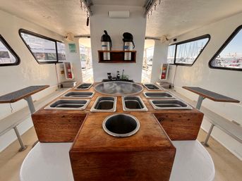 Interior of a fishing boat showing a wooden bait-prep station with multiple stainless steel bins, a round sink and cup hole, coffee carafes on a shelf, bench seating and marina views through side windows.