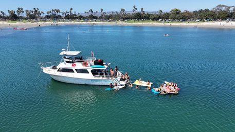 Charter boat anchored in a sunny coastal bay with people on inflatable rafts and paddleboards alongside, calm blue water and a sandy beach lined with palm trees in the background.