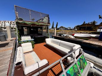 Houseboat docked at a sunny marina with a warm wood deck, white cushioned sofas, a shaded upper deck with netting, and nearby boats and waterfront homes under a clear blue sky.