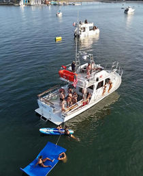 Summer boat party in a coastal harbor — group of friends on a white motorboat with a paddleboard and bright blue floating mat, sailboats and marina shoreline in the background