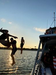 Silhouetted friends jumping off a boat into a sunlit bay at sunset with palm-tree lined shoreline and golden water reflections.