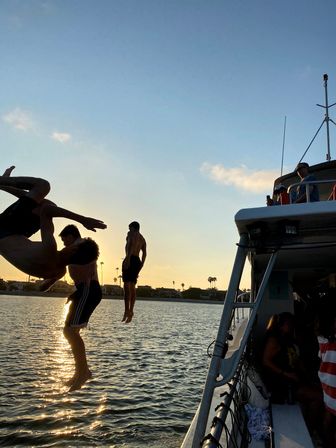Silhouetted friends jumping off a boat into a sunlit bay at sunset with palm-tree lined shoreline and golden water reflections.