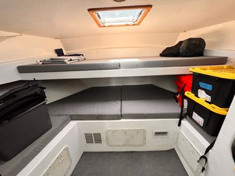 Cozy boat cabin V-berth with gray foam cushions on upper and lower bunks, overhead hatch letting in daylight, stacked yellow-lidded storage bins and red life jackets tucked to the side.