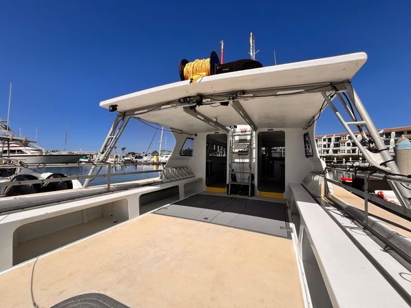 Open aft deck of a white boat docked at a sunny marina, ladder to the upper deck and a yellow rope reel on the roof against a clear blue sky