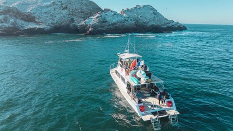 Sunlit tour boat carrying kayaks and people cruising turquoise ocean waters near a rugged rocky island shore.