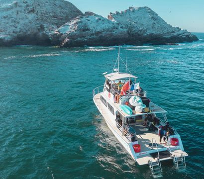 Sunlit tour boat carrying kayaks and people cruising turquoise ocean waters near a rugged rocky island shore.