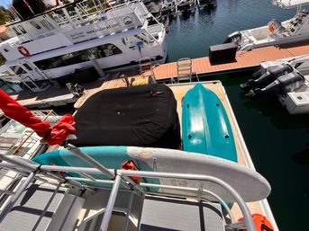 Top-down marina view of a boat deck with teal kayaks, a white paddleboard, black covered gear and moored passenger vessels along a wooden dock