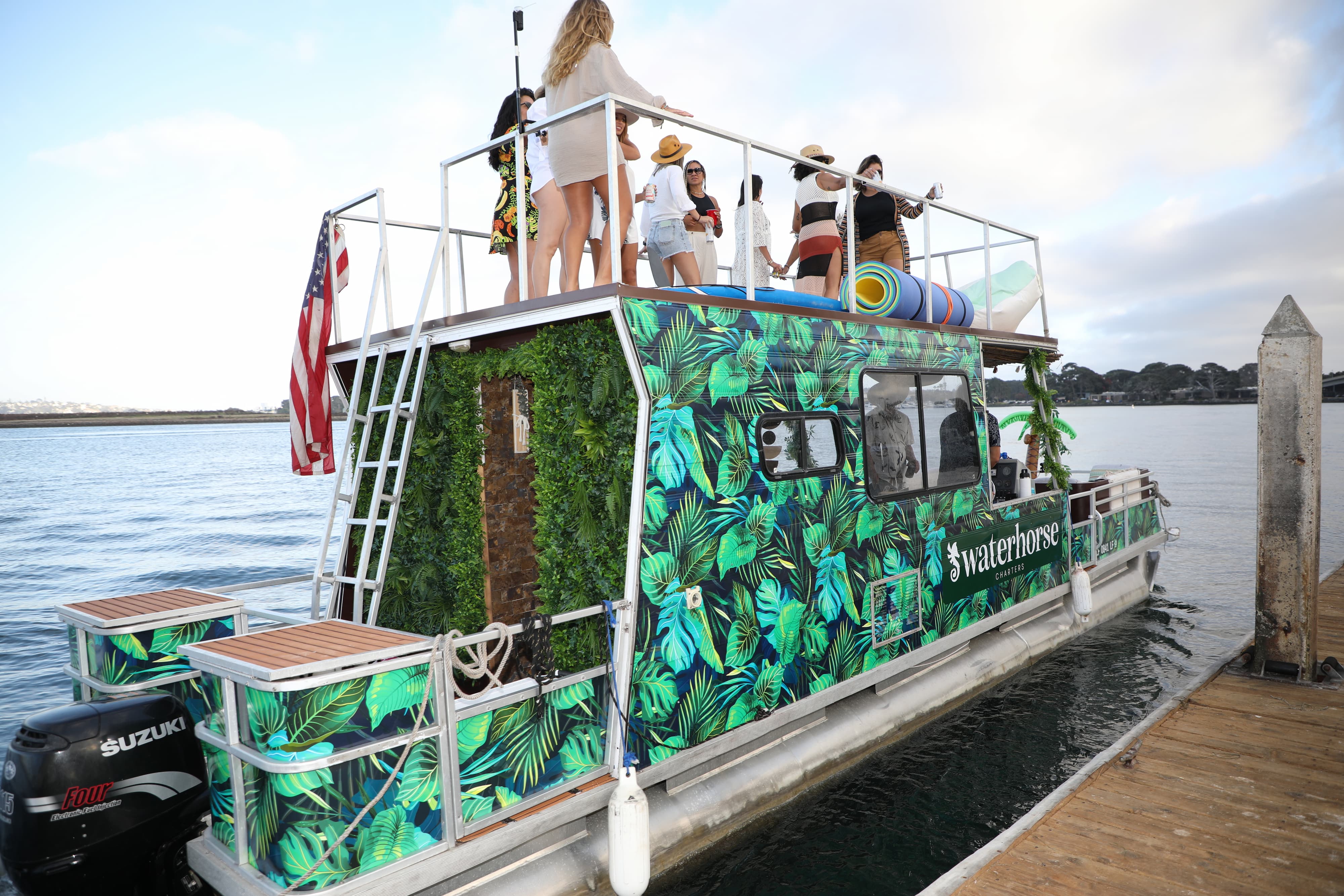 Tropical-leaf print party boat docked at a wooden marina pier, rooftop group of friends in summer outfits, American flag and calm bay under a cloudy sky.