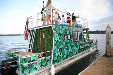 Tropical-leaf print party boat docked at a wooden marina pier, rooftop group of friends in summer outfits, American flag and calm bay under a cloudy sky.