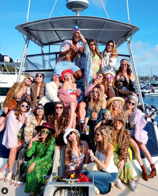 Large group of friends in colorful, boho-chic outfits and sunglasses posing and laughing on a sunny yacht deck during a lively boat party at a marina