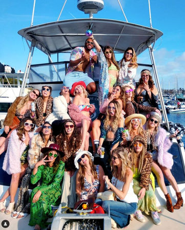 Large group of friends in colorful, boho-chic outfits and sunglasses posing and laughing on a sunny yacht deck during a lively boat party at a marina
