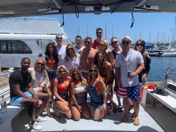 Group of friends posing on a sunny boat at a marina, wearing swimsuits and red-white-blue outfits and holding drinks for a festive summer yacht party