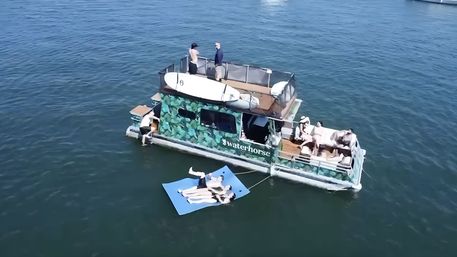 Camouflage-painted pontoon boat anchored in calm coastal waters on a sunny day, people lounging on the deck and on a tethered blue floating mat