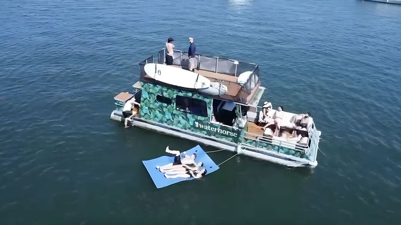 Camouflage-painted pontoon boat anchored in calm coastal waters on a sunny day, people lounging on the deck and on a tethered blue floating mat
