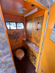 Compact boho-style orange portable restroom interior with tropical leaf stencil walls, small horizontal window, round tasseled toilet seat, narrow counter with fabric runner and jar, and wood-slat floor.