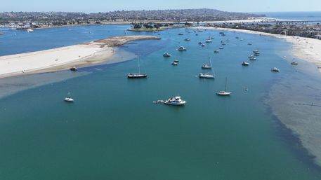 Aerial view of a turquoise coastal bay with anchored sailboats and yachts near a sandy barrier beach, distant shoreline and a pier.