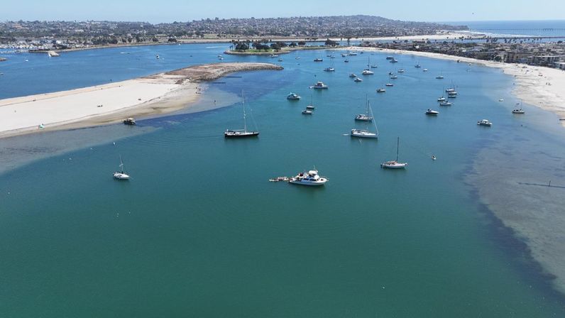 Aerial view of a turquoise coastal bay with anchored sailboats and yachts near a sandy barrier beach, distant shoreline and a pier.