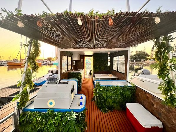 Tropical-themed pontoon boat at a marina during golden sunset, bamboo canopy with hanging tassels, lush green garlands, coolers and bench seating on a wooden deck, moored sailboats in the background.
